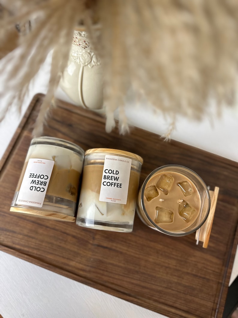 May include: Two glass candle jars with wooden lids and a glass of iced coffee on a wooden surface. The candles are labelled "Cold Brew Coffee" and the coffee is in a clear glass with ice cubes.