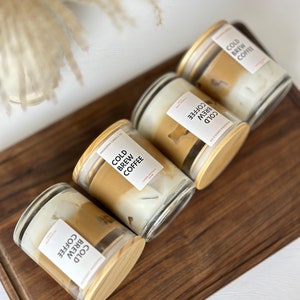 May include: Four glass candle jars with wooden lids. Each jar contains a candle with a layered design of brown and white wax. The labels on the jars read "Cold Brew Coffee".