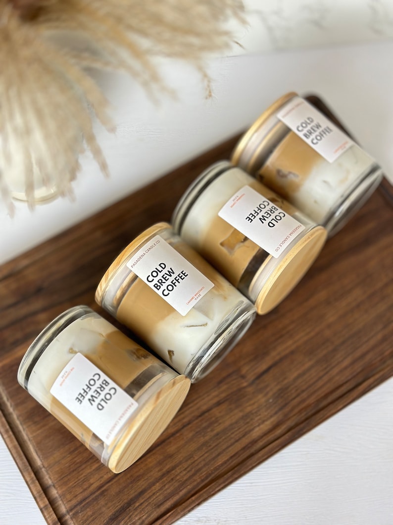 May include: Four glass candle jars with wooden lids. Each jar contains a candle with a layered design of brown and white wax. The labels on the jars read "Cold Brew Coffee".
