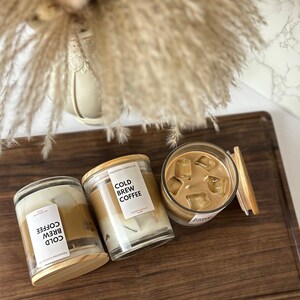 May include: Three glass candle jars with wooden lids. The candles are layered with a white wax base and a brown wax top. The labels on the jars read "Cold Brew Coffee".
