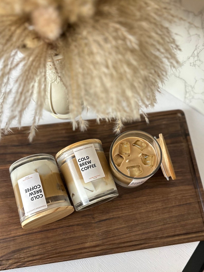 May include: Three glass candle jars with wooden lids. The candles are layered with a white wax base and a brown wax top. The labels on the jars read "Cold Brew Coffee".