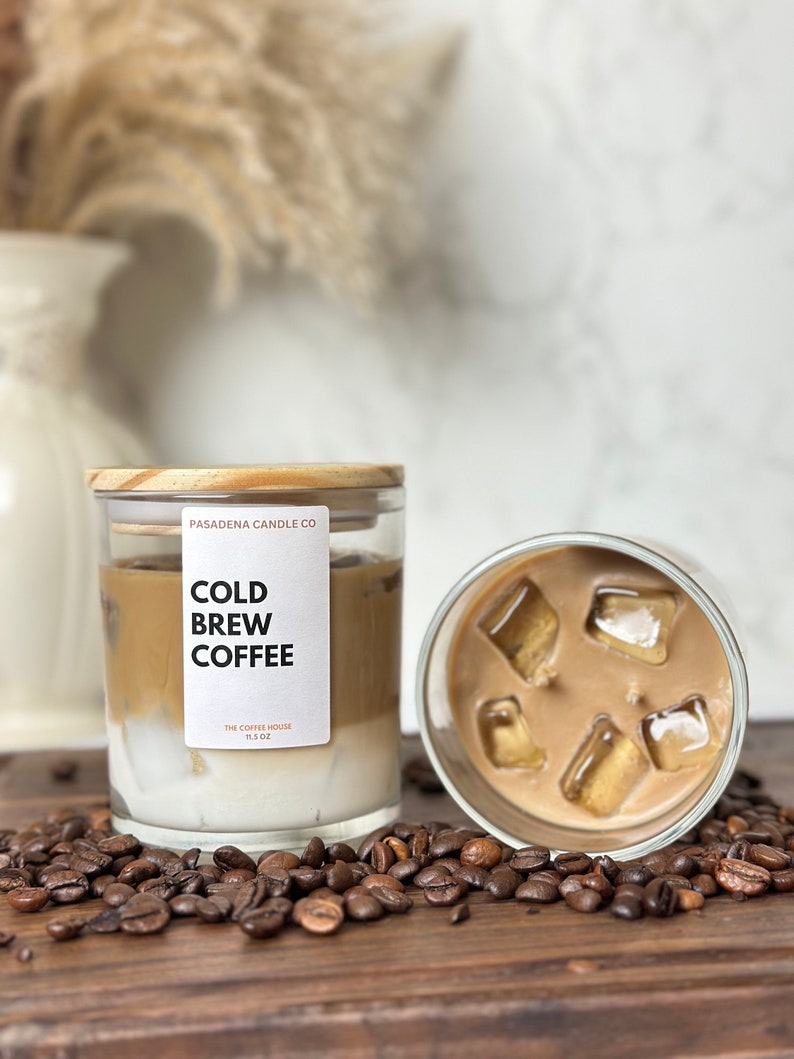 May include: A glass candle jar with a wooden lid. The candle is a layered scent of cold brew coffee with a white cream layer on top. The candle is surrounded by coffee beans. The label on the jar reads "PASADENA CANDLE CO. COLD BREW COFFEE THE COFFEE HOUSE 11.5 OZ."