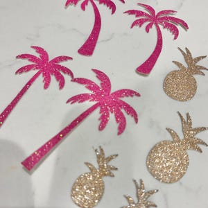 May include: Pink glitter palm tree and gold glitter pineapple cutouts. The tropical-themed decorations are scattered on a white surface. The palm trees have long trunks and detailed fronds. The pineapples have textured surfaces.