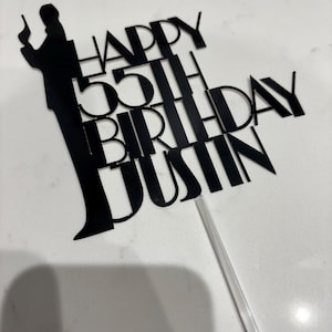 May include: Black cake topper with the text "HAPPY 55TH BIRTHDAY JUSTIN" and a silhouette of a person holding a gun. The topper is attached to a clear plastic stick for insertion into a cake.