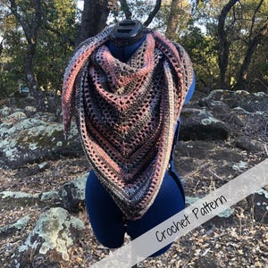 May include: A handmade crochet shawl in a triangle shape, displayed on a mannequin. The shawl features a mix of colors including pink, gray, and black. The text "Crochet Pattern" is visible on the image.