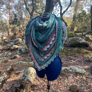 May include: A crocheted shawl in shades of teal, pink, and gray, displayed on a mannequin against a backdrop of rocks and trees.