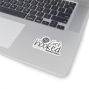 May include: A white sticker with the words "Get Hooked" in a handwritten font, with a yarn ball graphic. The sticker also includes the text "@jenetescreations". The sticker is placed on a silver laptop.