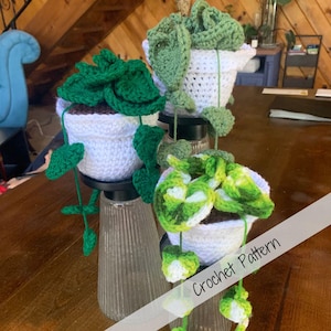 Puede incluir: Tres plantas en maceta de ganchillo con hojas verdes y blancas. Las plantas están dispuestas en una pantalla de varios niveles. El texto "Crochet Pattern" es visible en la esquina inferior derecha de la imagen.