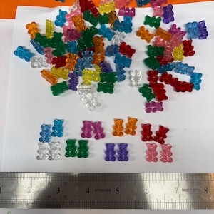 May include: A collection of colorful, translucent gummy bear-shaped beads in various colors, including blue, green, red, pink, and orange. A stainless steel ruler is placed below the beads.
