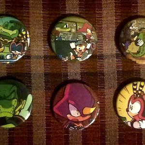 May include: Six round pinback buttons featuring colorful cartoon characters. The buttons display various characters in action poses, with a variety of colors including green, purple, yellow, and blue. The buttons are arranged on a brown striped surface.