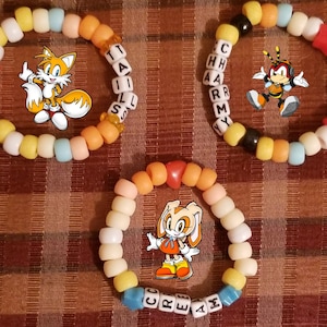May include: Three colorful beaded bracelets featuring cartoon characters. One bracelet has the name "TAILS" spelled out, another has "CHAMMY", and the third has "CREAM". The bracelets are made of various colored beads.