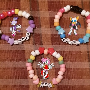 May include: Three colorful beaded bracelets with cartoon character images. One bracelet has the name "Blaze" spelled out with letter beads, another says "Rouge", and the third says "Amy". Each bracelet has star-shaped beads.