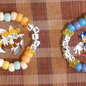 May include: Two colorful beaded bracelets featuring cartoon characters. One bracelet has the word "TAILS" spelled out with white letter beads and an image of Tails. The other bracelet has "SONIC" and an image of Sonic.