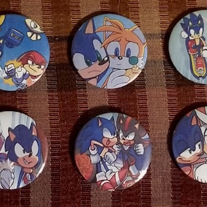 May include: A collection of six round pinback buttons featuring various characters from the Sonic the Hedgehog franchise. Each button displays a different pairing of characters in vibrant colors, set against a blue or white background.