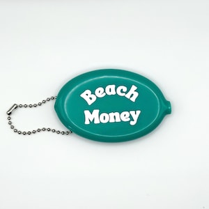 May include: Teal oval coin purse with the words "Beach Money" in white block letters. The purse has a silver chain attached to it. The background is white.
