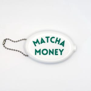 May include: White oval coin purse with the words "Matcha Money" in green lettering. The purse has a silver chain attached for easy carrying. The design is simple and modern, perfect for holding small change or other essentials.
