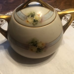 May include: A white porcelain sugar bowl with a gold lid and handles. The bowl is decorated with hand-painted floral designs in shades of yellow, green, and blue.