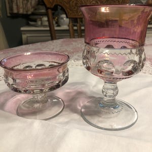 May include: Two antique clear glass goblets with a pink rim and base. The larger goblet has a tall stem and a wide bowl. The smaller goblet has a shallow bowl. Both have a decorative pattern.