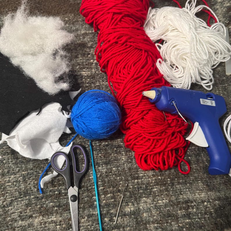 Puede incluir: Un proyecto de artesan&iacute;a con hilo rojo, blanco y azul, una bola de hilo azul, una pistola de pegamento, tijeras, un gancho de ganchillo y una aguja.