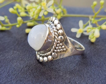 May include: A silver ring featuring a white moonstone set in an intricate, ornate design. The ring is displayed on a grey fabric background.