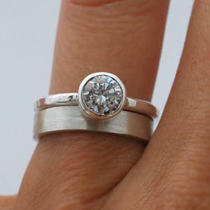 May include: A silver ring set with a clear, round gemstone in a bezel setting. The ring sits above a brushed silver band. The rings are shown on a finger against a soft, neutral background.