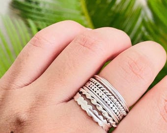 May include: A collection of silver rings with various textures and designs, including beaded, braided, and scalloped edges. The rings are stacked on a finger, showcasing the different styles. The background features green foliage.