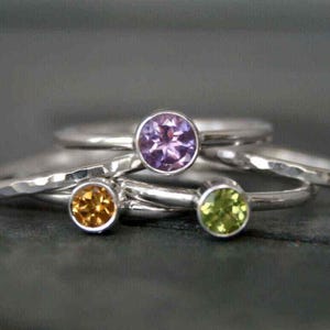 May include: A collection of silver rings with colorful gemstones. The rings feature a purple amethyst, a yellow citrine, and a green peridot. The gemstones are set in a simple bezel setting.
