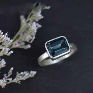 Puede incluir: Un anillo de plata con una piedra preciosa rectangular de color verde azulado. El anillo está sobre un fondo oscuro con una ramita de flores blancas y moradas. La piedra preciosa es de color verde azulado intenso.