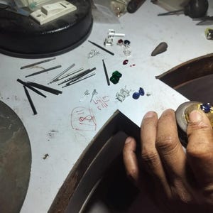May include: A jewelry maker's workbench with various tools and materials. Scattered across the surface are small silver earrings, gemstones in red, green, and blue, and several metal tools. The scene suggests jewelry design and craftsmanship.