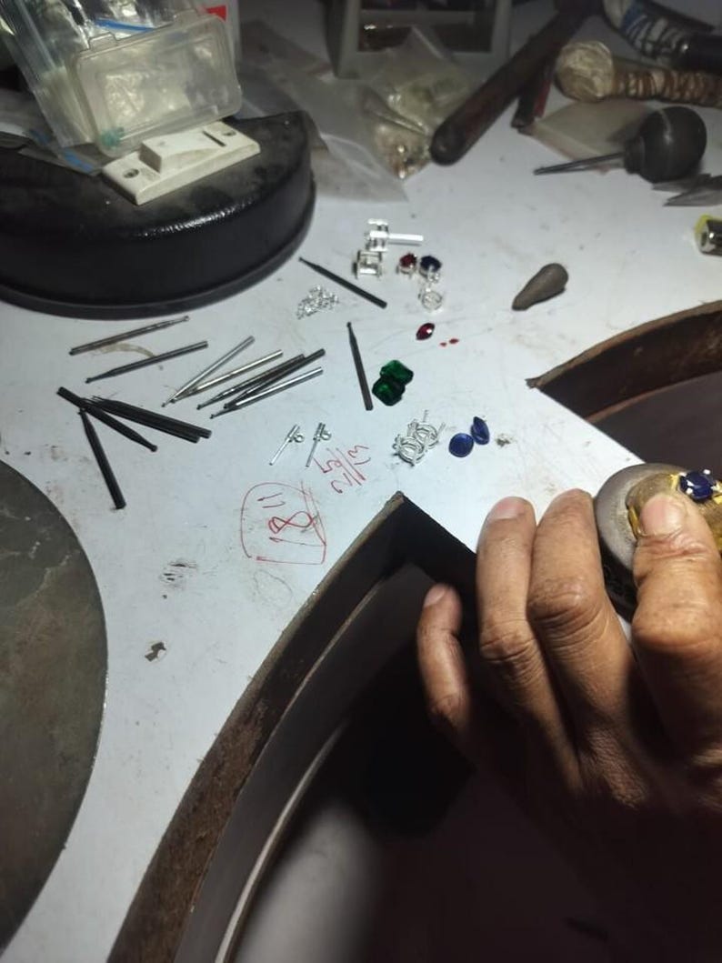 May include: A jewelry maker's workbench with various tools and materials. Scattered across the surface are small silver earrings, gemstones in red, green, and blue, and several metal tools. The scene suggests jewelry design and craftsmanship.