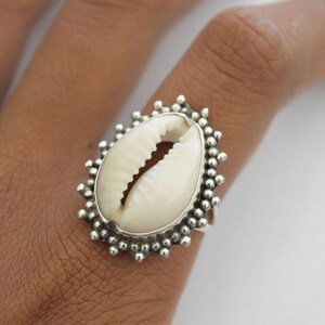 Könnte beinhalten: Ein Silberring mit einer großen, cremefarbenen Kaurischnecke als Mittelpunkt. Die Schale ist oval und in einen detaillierten, perlenbesetzten Silberrahmen gefasst. Der Ring wird an einem Finger getragen.