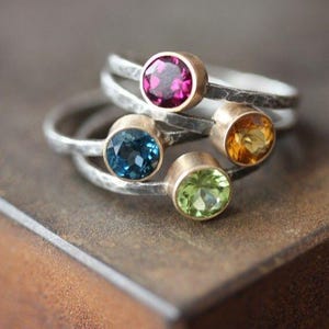 May include: A collection of gemstone rings with silver bands and gold bezels. The rings feature a variety of colorful gemstones, including pink, blue, yellow, and green. The rings are stacked on top of each other, creating a layered effect.