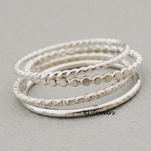 Puede incluir: Un conjunto de cuatro anillos plateados. Los anillos presentan diferentes texturas, incluyendo un diseño trenzado, un patrón de puntos, un acabado martillado y una banda lisa. Se muestran apilados sobre una superficie gris.