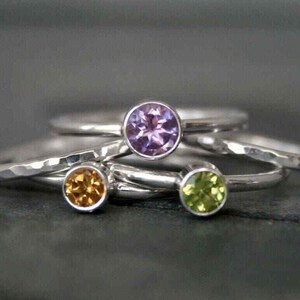 May include: Close-up of several silver rings with colorful gemstones. The rings feature a purple, yellow, and green gemstone. The rings are stacked on top of each other, with the gemstones facing up.