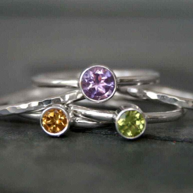 May include: Close-up of several silver rings with colorful gemstones. The rings feature a purple, yellow, and green gemstone. The rings are stacked on top of each other, with the gemstones facing up.