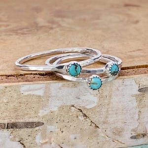 May include: Three silver rings with small, round turquoise stones. The rings are stacked on top of each other, with the turquoise stones visible. The rings have a hammered texture and are set against a light brown background.