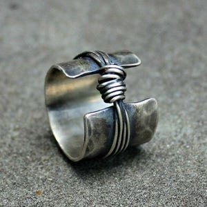 May include: A wide, silver ring with a hammered texture. The ring has a wrapped wire design in the centre, adding a unique, handcrafted element. The ring's design is open, with a gap in the band.