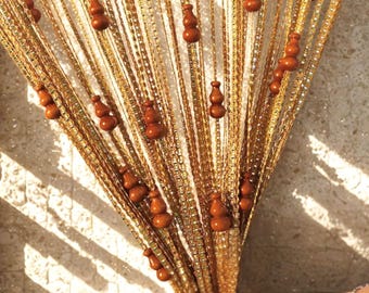 May include: A decorative beaded curtain with strands of gold-colored beads. Brown wooden gourd-shaped ornaments are evenly spaced along the strands. The curtain hangs in front of a light-colored wall, with shadows cast across it.