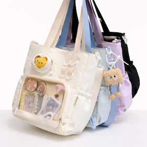 Puede incluir: Una colección de bolsos tote en varios colores pastel, incluyendo blanco, azul claro, lavanda y negro. Cada bolso tiene un compartimento principal, un bolsillo frontal transparente que muestra pequeños objetos y una correa a juego. Algunos bolsos tienen adornos decorativos.