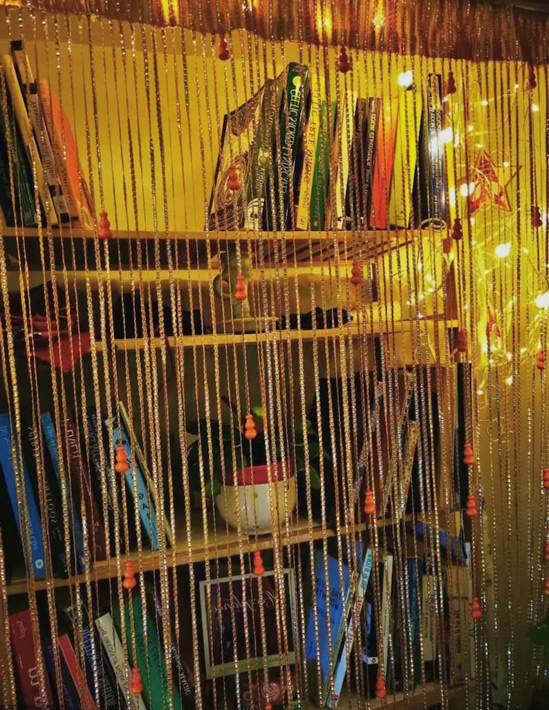 May include: A wooden bookshelf filled with books of various colors and sizes. The shelves are adorned with a beaded curtain, and the background is illuminated with warm lighting, creating a cozy atmosphere. A small potted plant sits on one of the shelves.