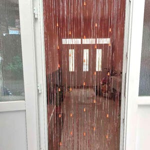 May include: A beaded curtain hangs in a doorway, featuring strands of reddish-brown beads with small orange accents. The curtain is framed by a white door and window frame, with a glimpse of the interior visible through the doorway.