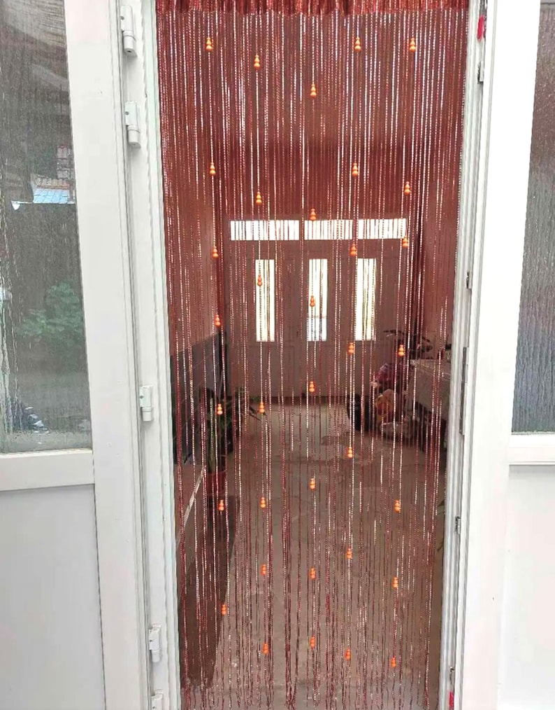 May include: A beaded curtain hangs in a doorway, featuring strands of reddish-brown beads with small orange accents. The curtain is framed by a white door and window frame, with a glimpse of the interior visible through the doorway.