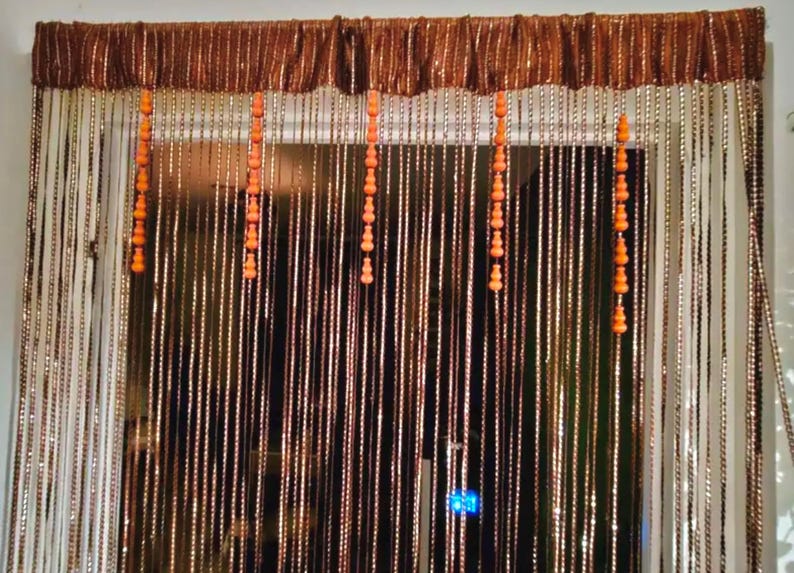 May include: A decorative beaded curtain with a brown valance. The curtain features long, vertical strands of beads in shades of brown and white, with orange beaded accents. The curtain is designed to hang in a doorway or window.