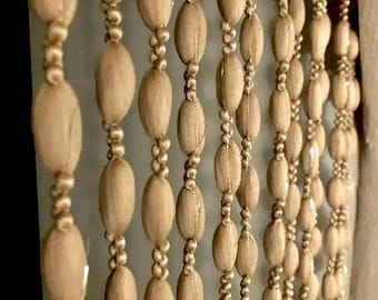 May include: Beaded curtain with rows of oval-shaped wooden beads in a light tan color. The beads are strung together with smaller, round beads, creating a decorative and textured effect. The curtain hangs in vertical lines.