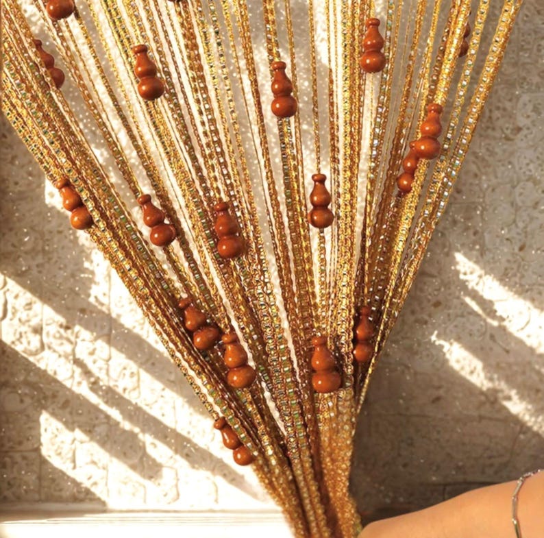 May include: A decorative beaded curtain with strands of gold-colored beads. Brown wooden gourd-shaped ornaments are evenly spaced along the strands. The curtain hangs in front of a light-colored wall, with shadows cast across it.