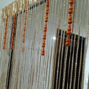 May include: A decorative beaded curtain with strands of clear beads and orange wooden accents. The top section features a gold-colored, textured fabric. The curtain hangs in front of a dark door frame.