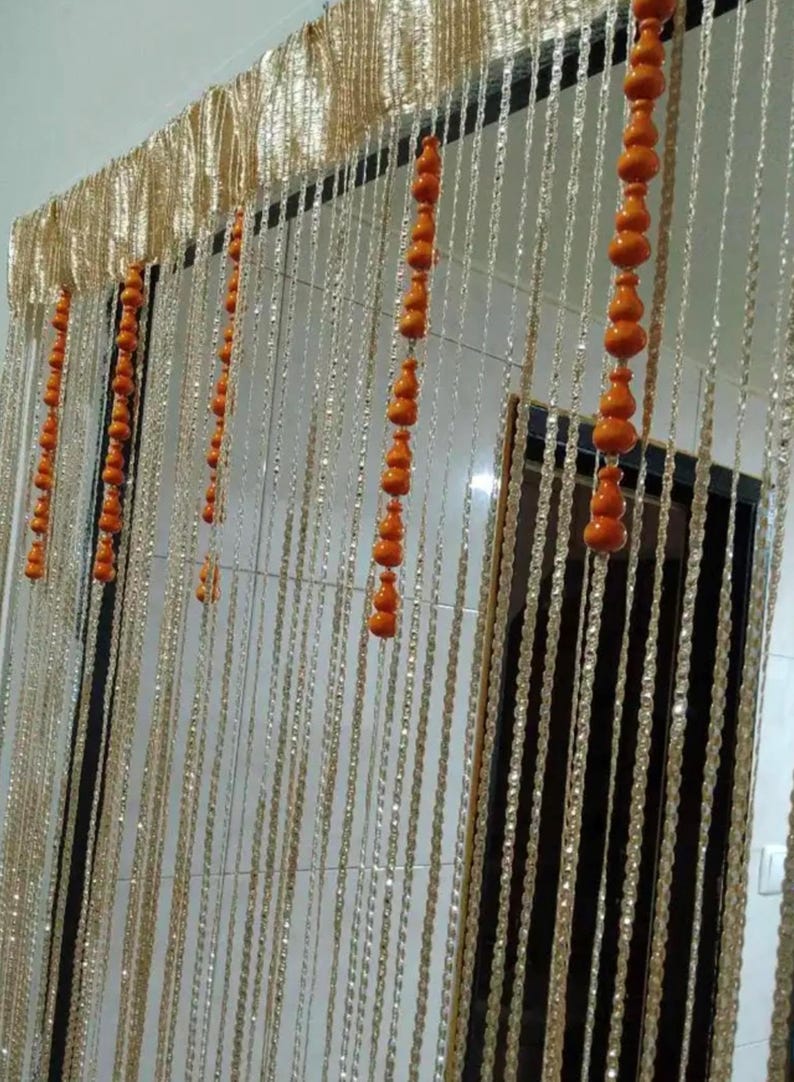 May include: A decorative beaded curtain with strands of clear beads and orange wooden accents. The top section features a gold-colored, textured fabric. The curtain hangs in front of a dark door frame.