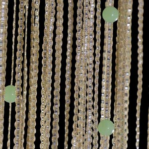 Pode incluir: Um close-up de uma cortina de contas decorativa. A cortina é feita de fios de pequenas contas retangulares transparentes. Espalhadas estão contas esféricas verde claro. O fundo é preto.