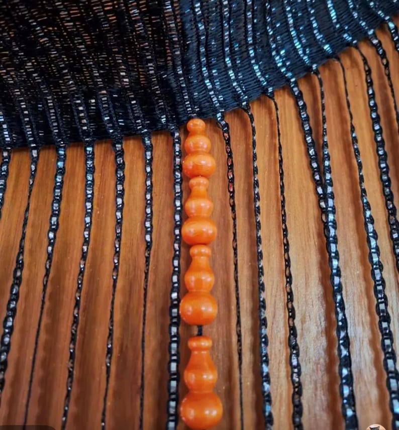 May include: An orange, decorative beaded curtain with a series of rounded shapes. The curtain hangs in front of a wooden surface. The top of the curtain is black with vertical strands of black and silver beads.
