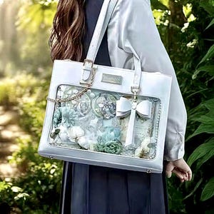 May include: A white tote bag with a clear front panel, displaying decorative items such as flowers, bows, and charms. The bag features a gold chain and a white bow. The bag is carried by a person.